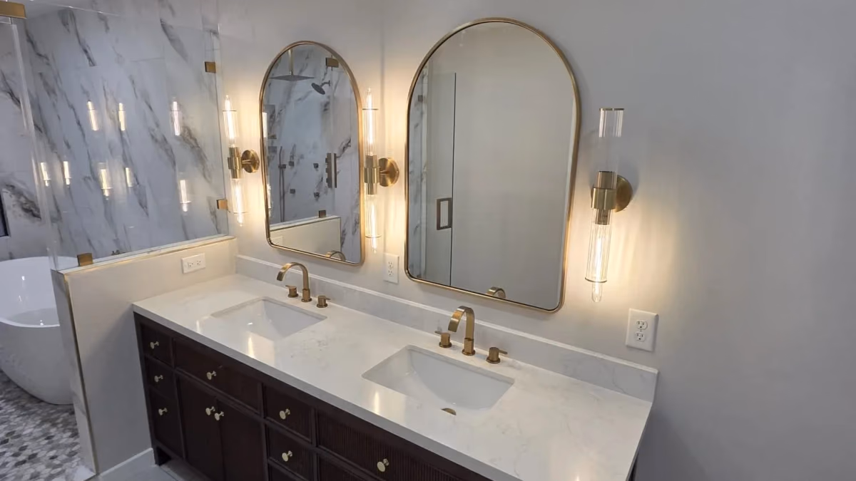 Double vanity with dark wood cabinetry, twin arched gold-framed mirrors, gold sconces, a white stone countertop, and marble-look tile
