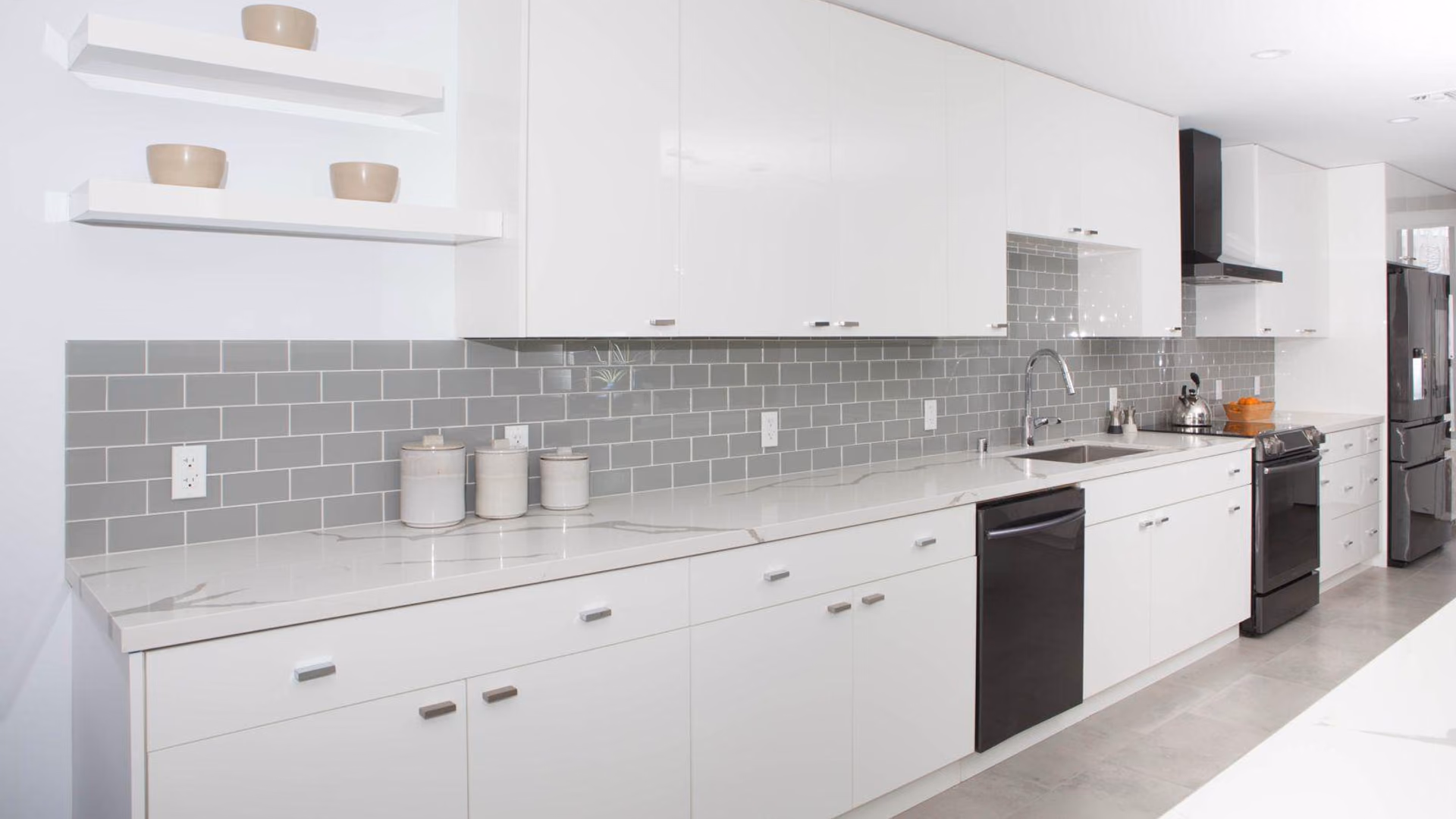 Completed kitchen remodel with white cabinetry, a gray glass subway backsplash, marble-look quartz countertops, floating shelves, and black appliances