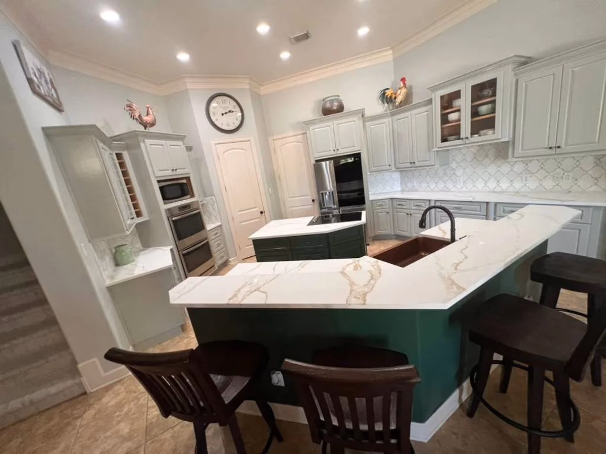 Sage-green kitchen island with a white quartz waterfall countertop and wood bar stools, set against white perimeter cabinetry with built-in wall ovens