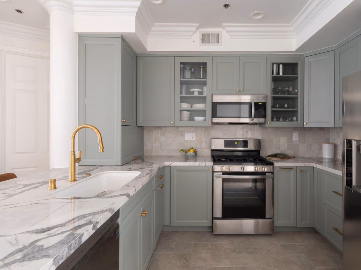 Sage-green kitchen with white marble countertops, a gold faucet, glass-front upper cabinets, and stainless appliances